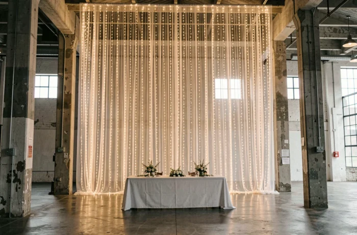 Guirlandes lumineuses en nappes verticales tombant du plafond d'un entrepôt reconverti en salle de mariage avec une table d'honneur en contrebas
